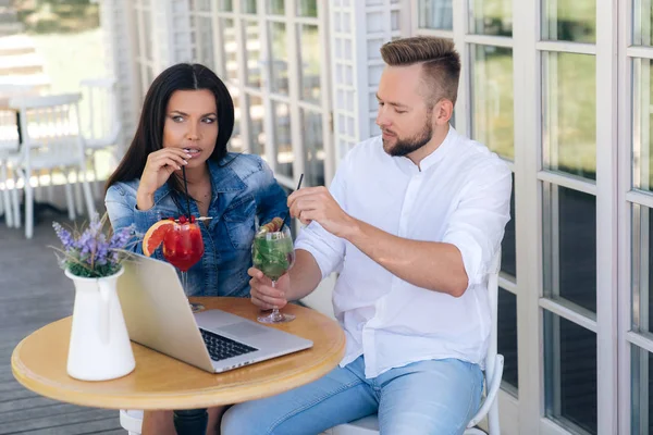 Freiberufler arbeiten in den Cafés, sie haben beschlossen, eine Pause einzulegen, sie haben Cocktails bestellt, das Mädchen nimmt nachdenklich einen Drink aus der Tube, der Mann erzählt ihr etwas, auf dem Tisch liegt ein Laptop.. — Stockfoto