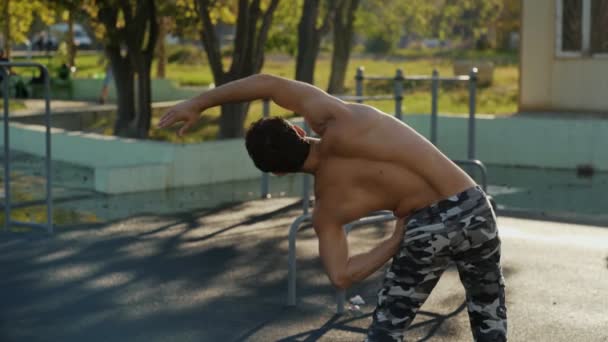 El atleta en el campo de deportes se calienta antes del entrenamiento. Un hombre musculoso va a practicar deportes al aire libre. El tipo con pantalones del ejército hace los ejercicios. Entrenamiento conceptual, crossfit, fitness, saludable . — Vídeo de stock