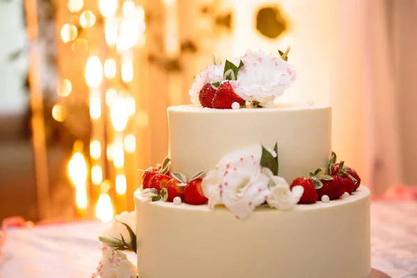 Крупним планом на столі лежить весільний білий вершковий торт. Десерт прикрашений ягодами стиглої червоної полуниці і живими білими квітами. Ідеї для прикраси весільного торта, стиль весілля . — стокове фото