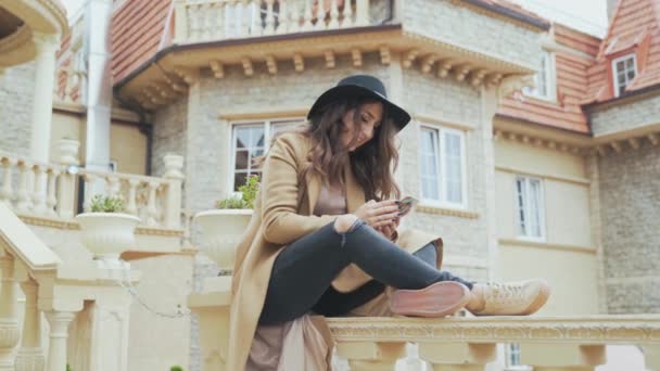 Atractiva europea sonriente con el pelo rizado, con un sombrero y un abrigo elegantes, hablando con alguien en su teléfono inteligente, haciendo una cita, escribiendo SMS desde su teléfono . — Vídeo de stock