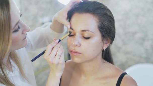 Närbild på en vacker ung brunett kom till hennes makeupartist att göra en dag make-up. En makeup specialist gäller en concealer klienter övre ögonlocket med en borste. Slow motion. — Stockvideo