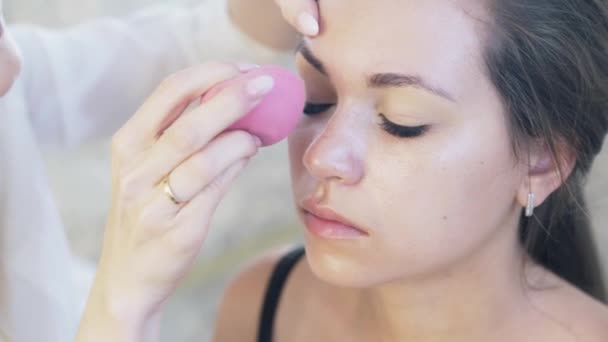 Nahaufnahme beschnittene Rahmen junger Frauen kamen zum Make-up. Der Visagist interpretiert Concealer auf dem Oberlid zum Klienten, bläst ihn mit Hilfe kleiner Paralonkugeln auf. Zeitlupe. — Stockvideo