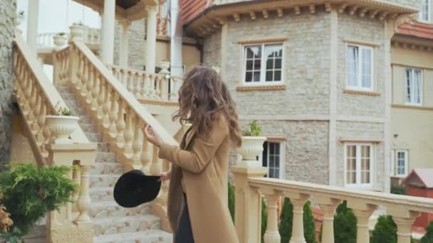 Une jolie jeune femme se promène dans le château, admire les anciens escaliers et tours, porte des vêtements élégants, porte un manteau et un chapeau, a les cheveux longs bouclés, tourne, sourit, bénéficie d'une visite guidée . — Video