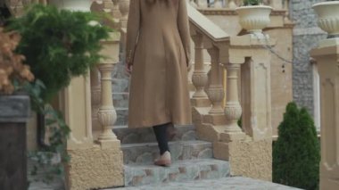 Silah alt, yakın çekim, yavaş açı, siyah şık bir şapka giyen bir uzun kaşmir ceket giymiş genç bir kız bir ev adımlarla tırmanıyor.