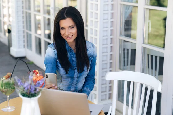 Attraktive junge Mädchen sitzt in einem Café, liest SMS auf ihrem Smartphone und lächelt, nutzt Laptop für die Arbeit. das Konzept der Geräte, der Technologie, des Internets. — Stockfoto