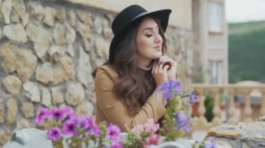 Yakın çekim Genç kadın Avrupa görünümü, kıvırcık siyah saçlı, şapka ve sonbahar bir ceket giyer, onların taş balkonda duran, çiçeklerle süslenmiş, yabancı bir şehirde sokak hayran.