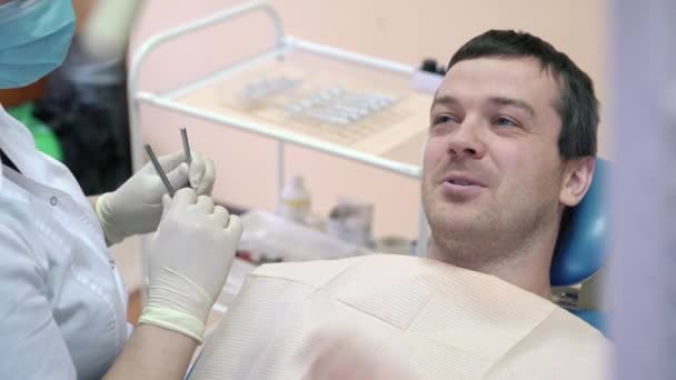 Un paciente masculino está acostado en una silla dental en una clínica y hablando con un médico, sonriendo y preparándose para los procedimientos. Consulta en el consultorio dental — Vídeos de Stock