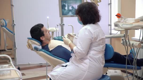 En manlig patient är liggande på en stol i en klinik och får samråd från en tandläkare. Läkaren undersöker tänderna och förbereder sig för att utföra de förfaranden och behandling. — Stockvideo