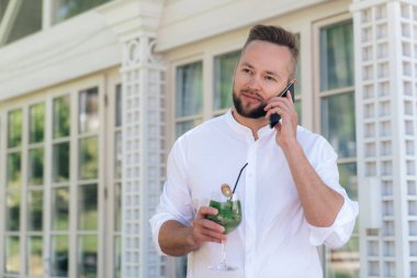 Şık ve çekici bir genç adam boyunca Teras, bir kokteyl yürüyüşler ile birine gülümsüyor telefonda konuşuyor. Kısa saç kesimi ve düzgün sakalı beyaz gömlekli olan yakışıklı bir adam görüşmeler olduğunu.