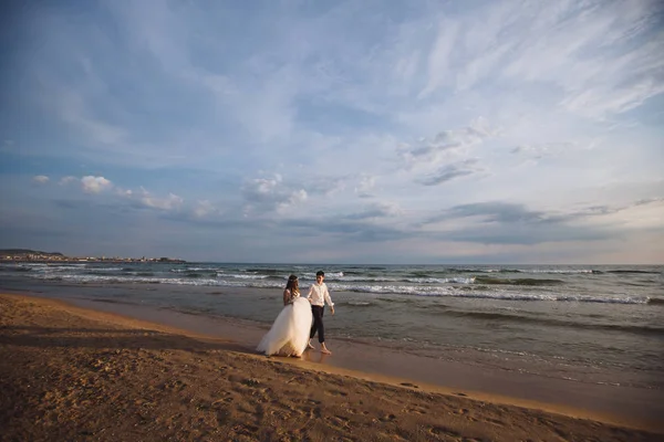 Una bella coppia di sposi novelli, gli sposi che camminano sulla spiaggia. Splendido tramonto e cielo. Abiti da sposa, un abito bianco di lusso per una ragazza. Concetto di famiglia, luna di miele . — Foto Stock