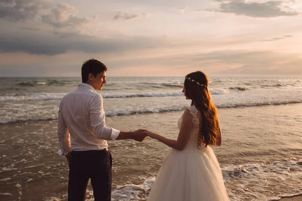 Una bella coppia di sposi novelli, gli sposi che camminano sulla spiaggia. Splendido tramonto e cielo. Abiti da sposa, un abito bianco di lusso per una ragazza. Concetto di famiglia, luna di miele . — Foto Stock