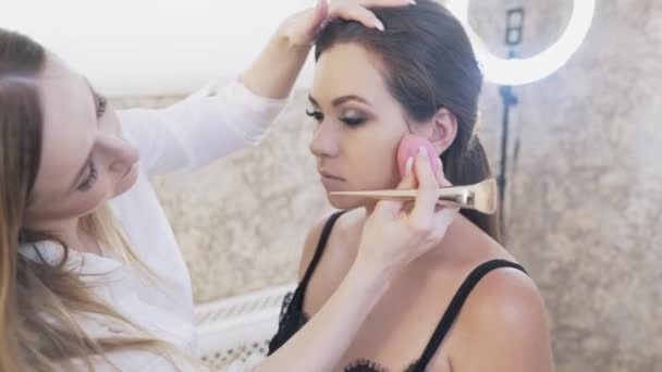 Zbliżenie: Knaga piękności siedzi na krześle z make-up artist z makijaż smoky eyes, popycha dziewczyna modele skóry z gąbką, koryguje błędy w makijażu. — Wideo stockowe