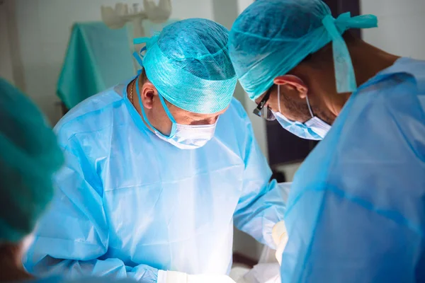 Two doctors in sterile medical clothes perform an operation in a hospital. Professional work in the clinic, surgeons are men with an assistant woman. The concept of health, medicine. — Stock Photo, Image