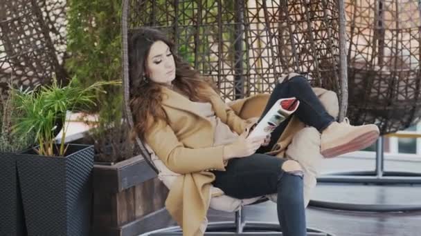 Ein schönes Mädchen in Pullover und Mantel sitzt auf einem Stuhl in einem Café und telefoniert emotional. Frau löst geschäftliche Probleme und hält eine Zeitschrift — Stockvideo