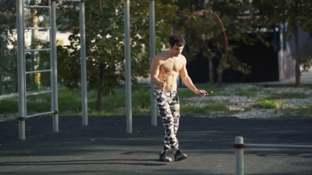 In Zeitlupe springt ein Athlet in Camouflage-Hosen mit einem Springseil über den Sportplatz. Muskelprotz wärmt sich auf, bevor er Sport treibt. Fitness-Konzept, Gesundheit. — Stockvideo