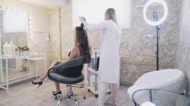 Model makyaj sanatçısı sandalyede oturur, stilist saçlarını twirls bukleler forseps üzerinde fırçalar. Siyah elbiseli bir kadın makyaj ve saç yapmak için güzellik salonuna geldi.