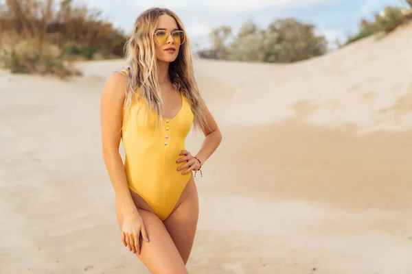 Sexy Model in gelbem Badeanzug und Sonnenbrille posiert am weißen Sandstrand. — Stockfoto