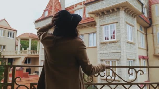 Una chica alegre en un abrigo largo lleva un sombrero negro con estilo por primera vez llegó a Europa, agita la mano a los transeúntes, se para en el puente, admira los edificios antiguos — Vídeo de stock