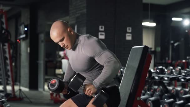 Close-up homem musculoso forte está treinando no ginásio escuro. O atleta senta-se em um banco e pressiona halteres, treina ombros e braços. Um cara esportivo está envolvido em fitness. Conceito desporto e saúde . — Vídeo de Stock