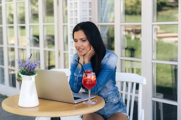 Ein attraktives Mädchen sitzt an einem Tisch in einem Café, trinkt einen roten Cocktail und schaut interessiert in den Laptop. Konzept der Technologie, Gadget, Menschen, Freelancing. — Stockfoto