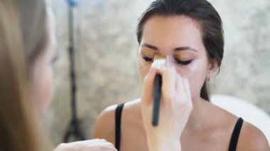 Genç bir sarışın sarışın makyöz Close-up bir genç esmer kız boyar, bir kapatıcı yüzünü geniş bir fırça ile uygulanır. Kavramı kişi, makyaj güzellik sektöründe çalışıyor. Ağır çekim.