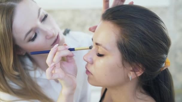 Krásná mladá Bruneta detail přišel její maskérka udělat denní make-up. Specialista na make-up platí korektor na horní víčko klienty pomocí štětce. — Stock video