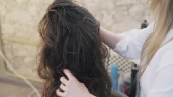 Nahaufnahme, beschnittener Rahmen, schönes lächelndes junges Mädchen mit Abendschminke, das auf dem Stuhl eines Friseurs sitzt, sich die Haare macht, die Haare kraust. Stylist Hände formen die Haare. — Stockvideo