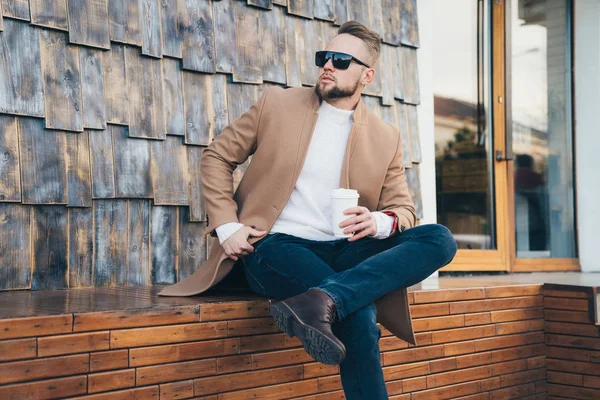 Hombre guapo con ropa elegante sentado cerca del café con las piernas cruzadas — Foto de Stock