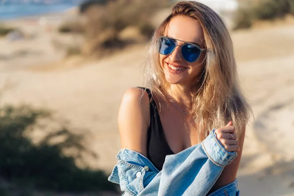 Närbild porträtt av vackra träning blond modell i svart baddräkt och solglasögon poserar på stranden. En flicka med en sexig bild håller en jeansjacka i händerna, sola på semester. — Stockfoto