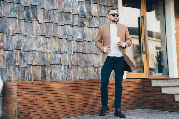 Attraktiver bärtiger Mann mit braunem Mantel, Sonnenbrille und Kaffeetasse in der Hand im Caféhintergrund stehend, Kopierraum auf Steinmauer. — Stockfoto