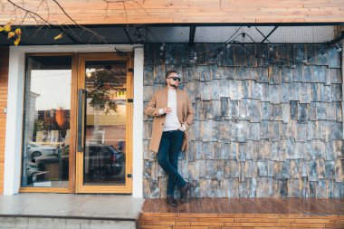 İşadamı kahverengi ceket, güneş gözlüğü ve kahve Cafe arka plan, taş duvar üzerinde kopya alanı poz onun elinde.