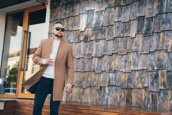 Lifestyle Mode Porträt eines jungen europäischen Mannes in beigem Mantel und Tasse Kaffee, Kopierraum auf Steinfliesen Wand. — Stockfoto