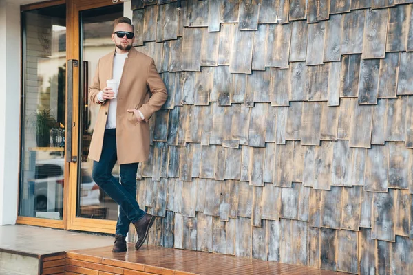 Empresario en abrigo marrón, gafas de sol y taza de café en sus manos posando sobre el fondo de la cafetería, espacio de copia en la pared de piedra . — Foto de Stock