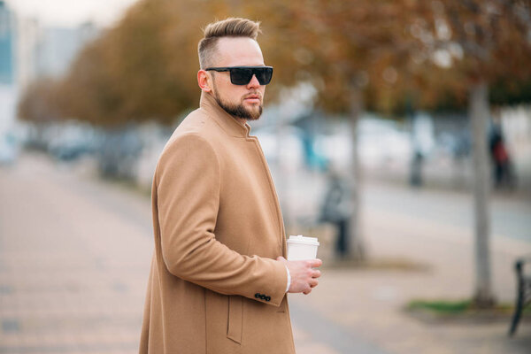 Handsome bearded young man in sunglasses drinking coffee in park, copy space on cup