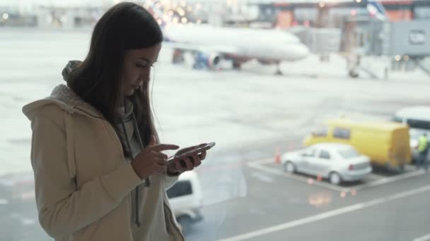 Aantrekkelijk meisje permanent bij raam in de terminal van de luchthaven en met behulp van de telefoon in de achtergrond van vliegtuig. — Stockvideo