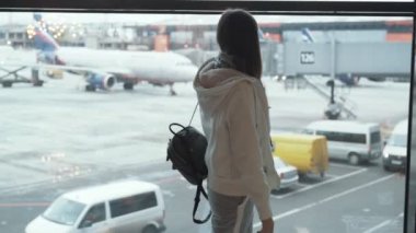 Arka görünümü kız sırt çantası ile uçak pencereden dışarı bakar ve elini dalgalar. Havaalanı terminal penceresinde, kadın ayakta. Kavram seyahat, Tur, uçuş, savaş uçağı, gelenler, lounge, pist, check-in