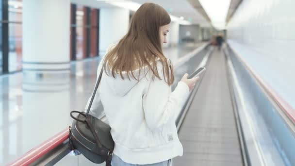 国際空港の動く歩道上で携帯電話上に裏面ビュー ヨーロッパ女性テキスト メッセージ. — ストック動画