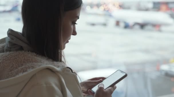 Vista lateral morena mujer usando teléfono inteligente cerca de la ventana del aeropuerto . — Vídeos de Stock