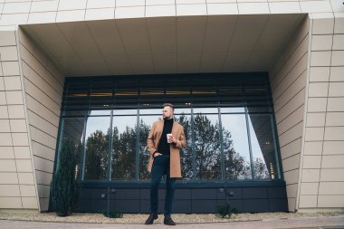 Çekici bir adam trendy kahverengi ceket ve iş merkezine yakın poz elinde kahve
