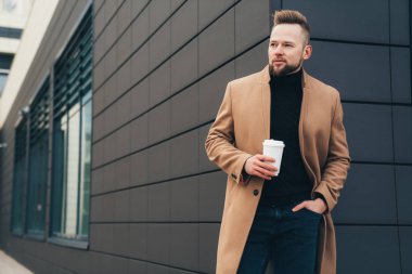 Çekici bir adam trendy kahverengi ceket ve iş merkezine yakın poz elinde kahve