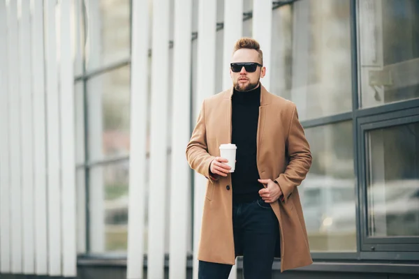 Schöner bärtiger junger Mann mit Sonnenbrille, der eine Tasse Kaffee zum Mitnehmen in der Nähe des Geschäftszentrums hält. — Stockfoto