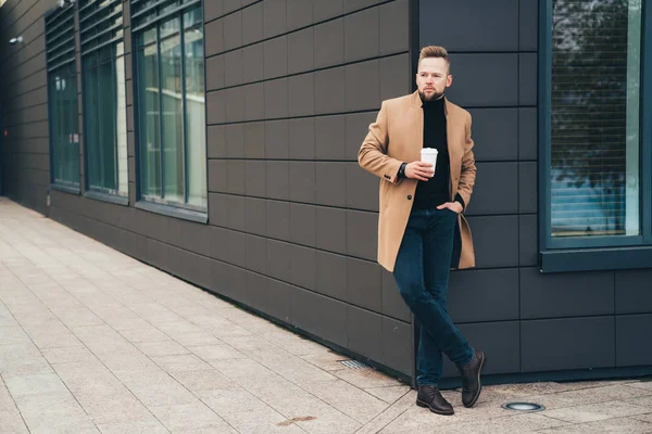 Attraktiver Mann im trendigen braunen Mantel und Kaffee in der Hand, der in der Nähe des Business Centers posiert — Stockfoto