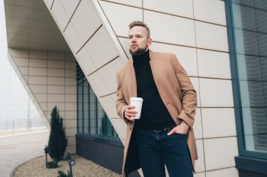 Çekici bir adam trendy kahverengi ceket ve iş merkezine yakın poz elinde kahve