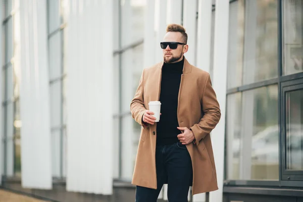 Schöner bärtiger junger Mann mit Sonnenbrille, der eine Tasse Kaffee zum Mitnehmen in der Nähe des Geschäftszentrums hält. — Stockfoto