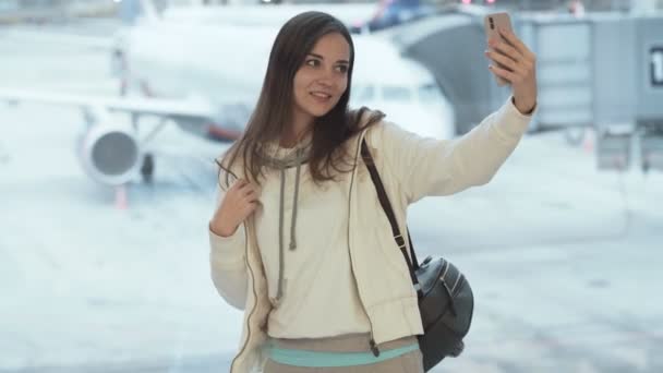 Дівчина робить селфі, показуючи великий палець вгору жестом і брязкальцями біля вікна в аеропорту — стокове відео