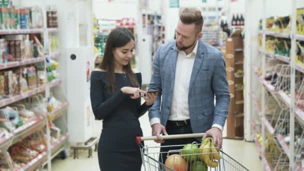 Kadın smartphone ürün ve adam listesi meyve seçer ve süpermarkette kokuyor denetlemek için kullanır — Stok video