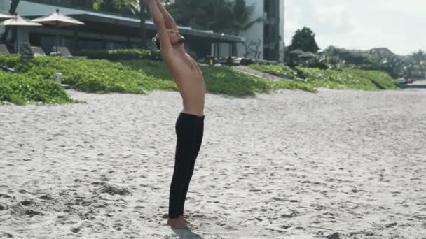 Hombre de pie en pose de yoga y haciendo ejercicios de estiramiento en la playa de arena, steadicam shot — Vídeo de stock