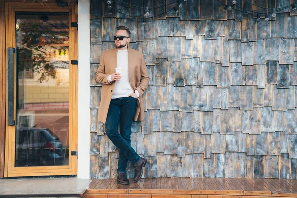 Geschäftsmann in braunem Mantel, Sonnenbrille und Kaffeetasse in der Hand posiert auf Café-Hintergrund, Kopierraum an Steinmauer. — Stockfoto