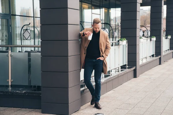 Hombre atractivo en abrigo marrón de moda y café en su mano posando cerca del centro de negocios — Foto de Stock