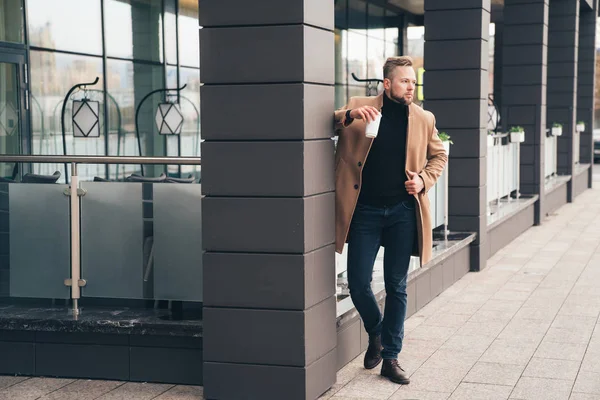 Attraktiver Mann im trendigen braunen Mantel und Kaffee in der Hand, der in der Nähe des Business Centers posiert — Stockfoto
